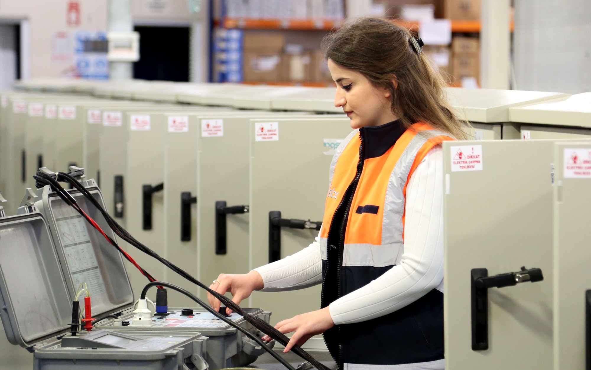 Woman electrician in a safety vest testing equipment beside electrical control panels